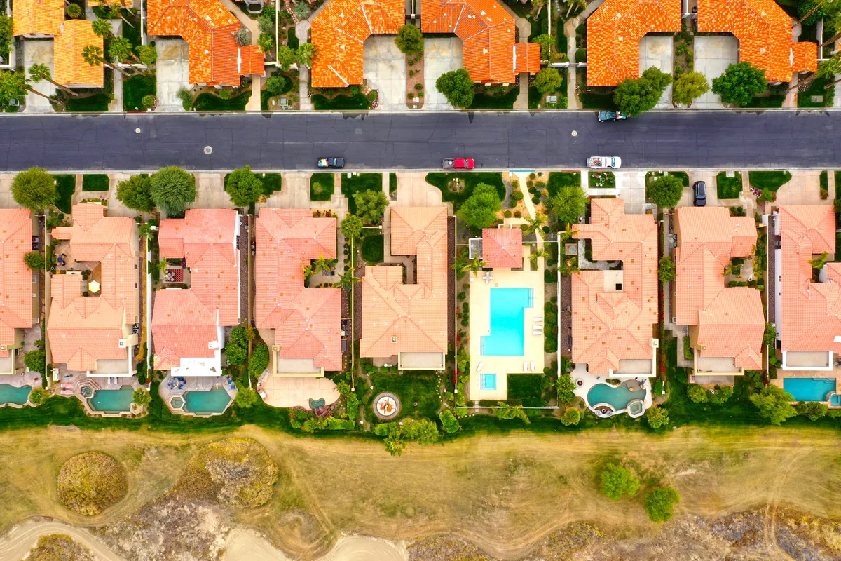 Aerial view of a row of tile-roof homes with private backyard pools beside open desert