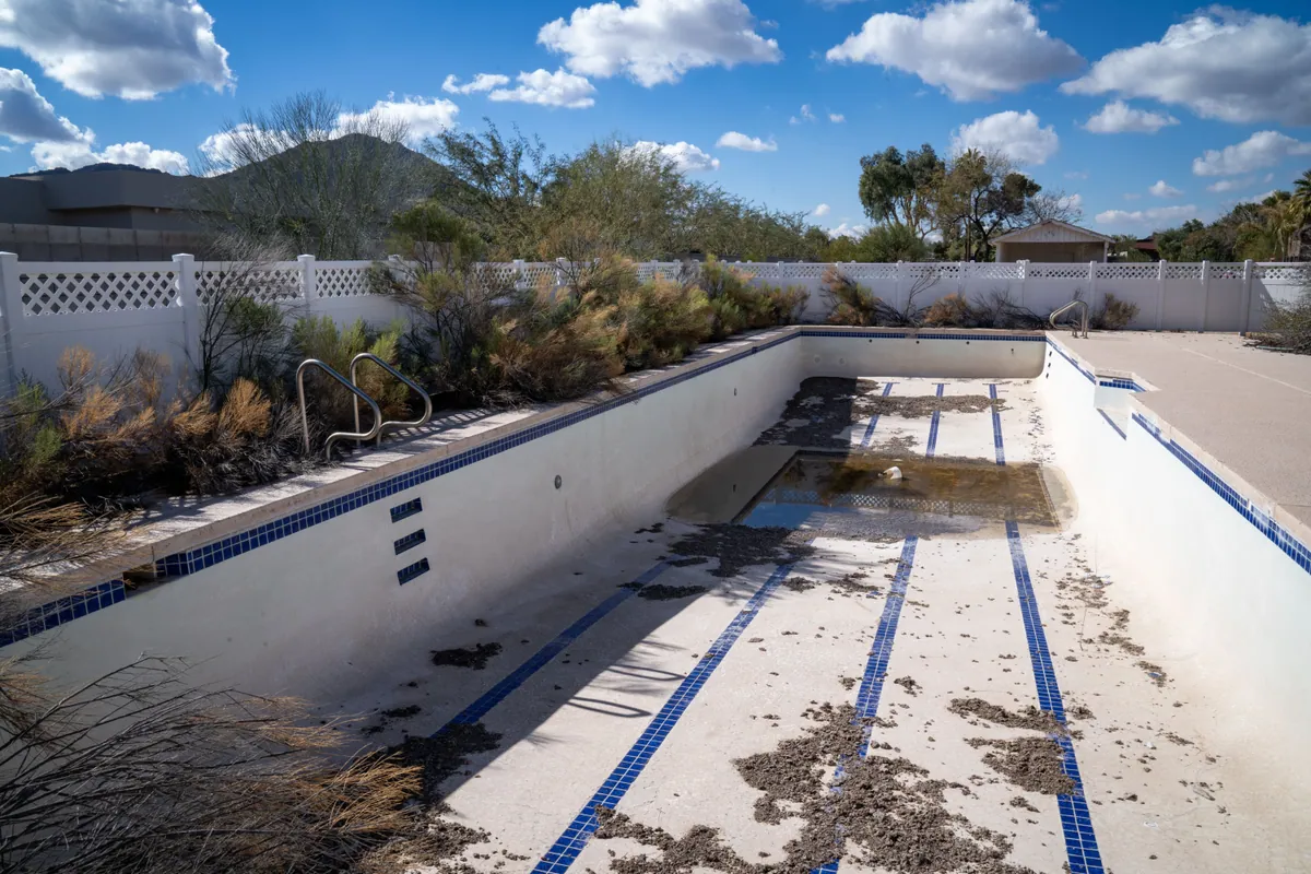 Drained pool with stained, worn plaster and debris on the floor, desert mountains beyond the fence